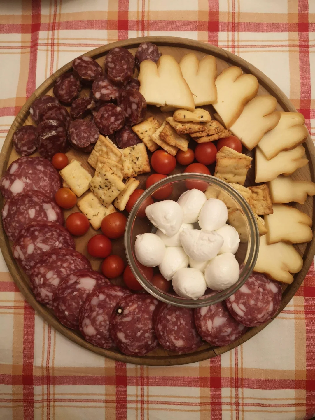Round wooden platter with sliced cured meats, cherry tomatoes, crackers, sliced cheese, and a bowl of mozzarella balls, on a red and white checkered tablecloth.