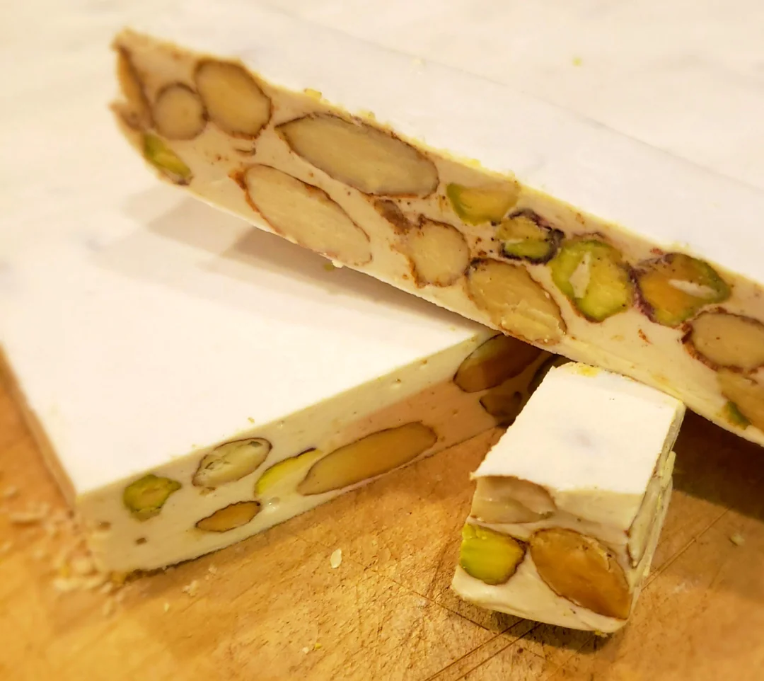 A close-up of nougat bars filled with whole almonds and pistachios, stacked on a wooden surface. The nougat is creamy white with visible nut pieces throughout.