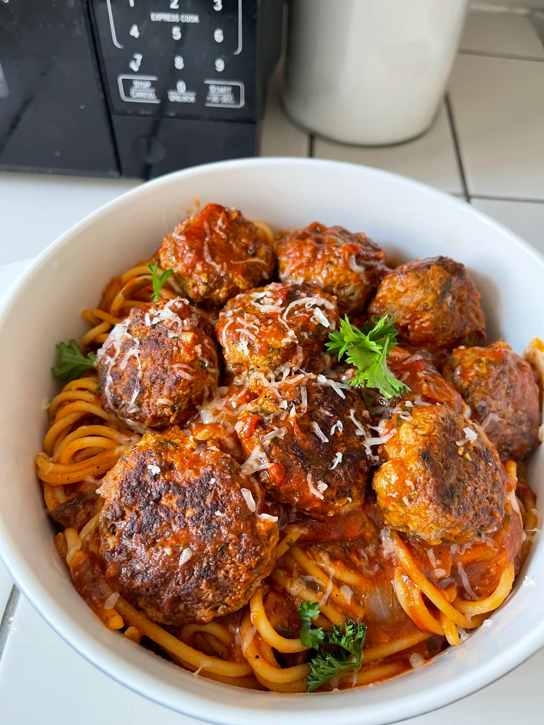 A white bowl filled with spaghetti and meatballs topped with tomato sauce, grated cheese, and parsley sits on a kitchen counter near an appliance.