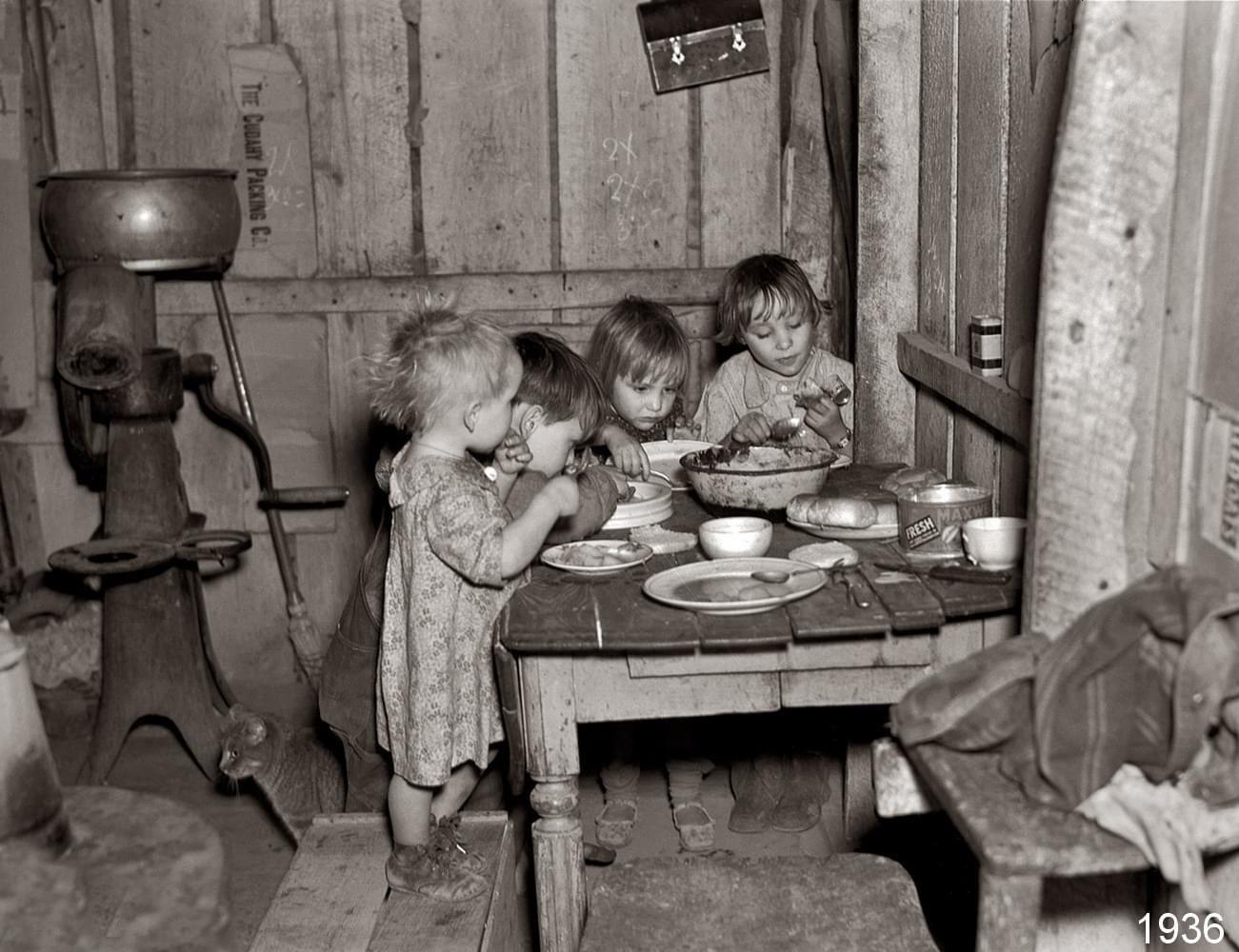 Four young children in worn clothes eat at a small wooden table in a rustic, shabby room, evoking the simplicity of nostalgic dinners from the past. A metal grinder and basic kitchenware are visible. Photo is in black and white, dated 1936.