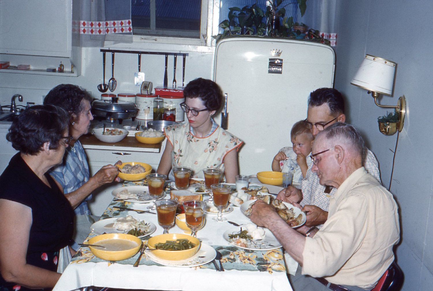 Six adults and one child sit around a kitchen table sharing a meal, evoking the warmth of nostalgic dinners. The table is set with plates of food and glasses of iced tea, while a refrigerator and kitchenware are visible in the background.