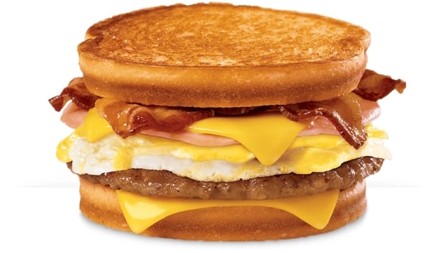 A breakfast sandwich with sausage, egg, bacon, ham, and melted cheese between two slices of golden toasted bread.