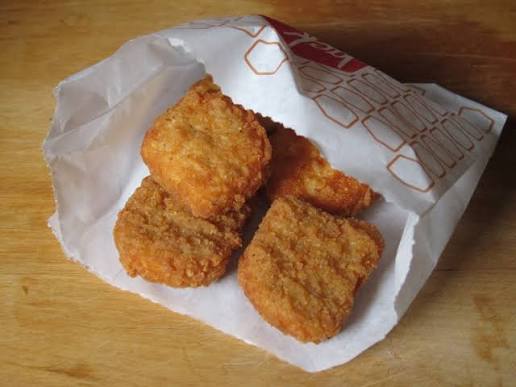 Four crispy chicken nuggets in a white paper bag, partially open, resting on a wooden surface.