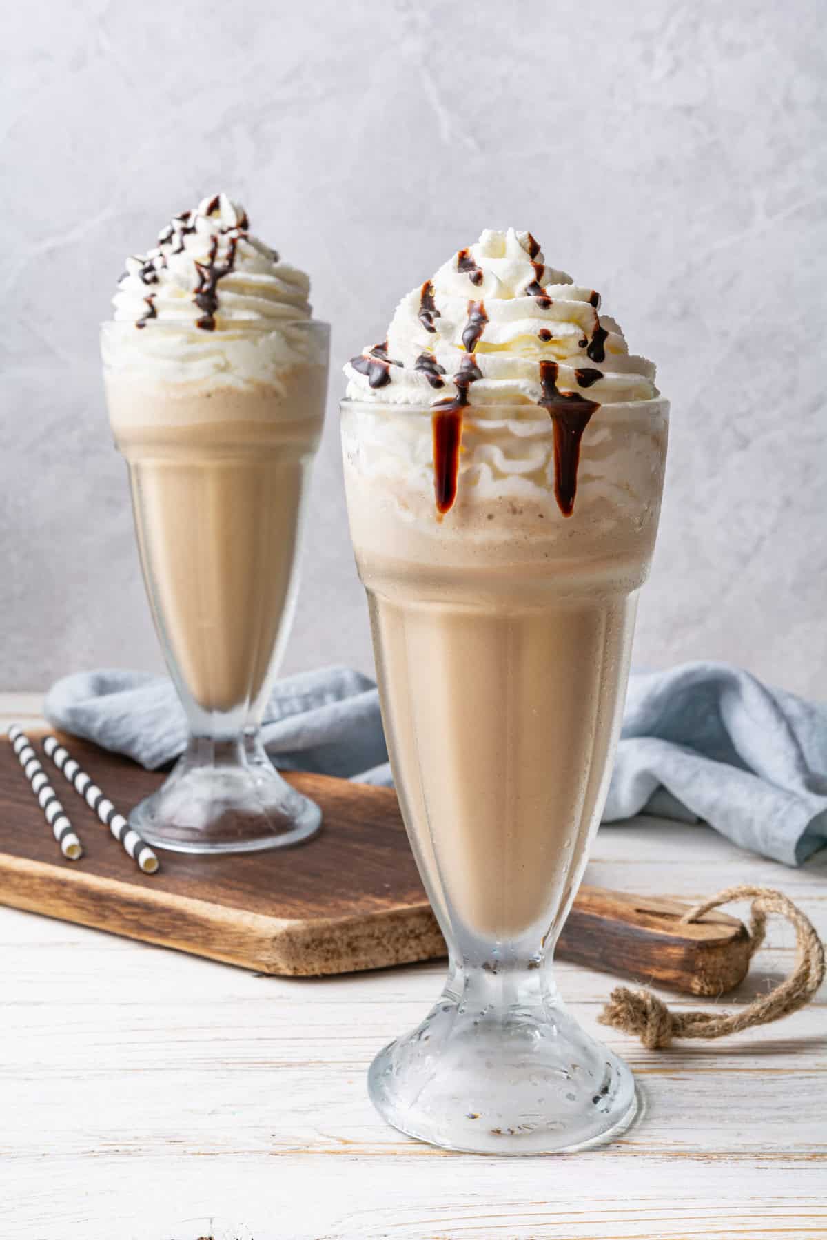 Two iced coffee drinks in tall glasses topped with whipped cream and chocolate syrup, placed on a wooden board with striped straws nearby, set on a white table.