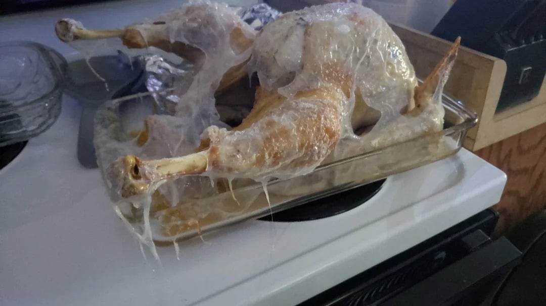 A whole raw turkey covered in a layer of plastic wrap sits in a glass baking dish on a white stovetop. The plastic is clinging to the meat and stretched across the surface.