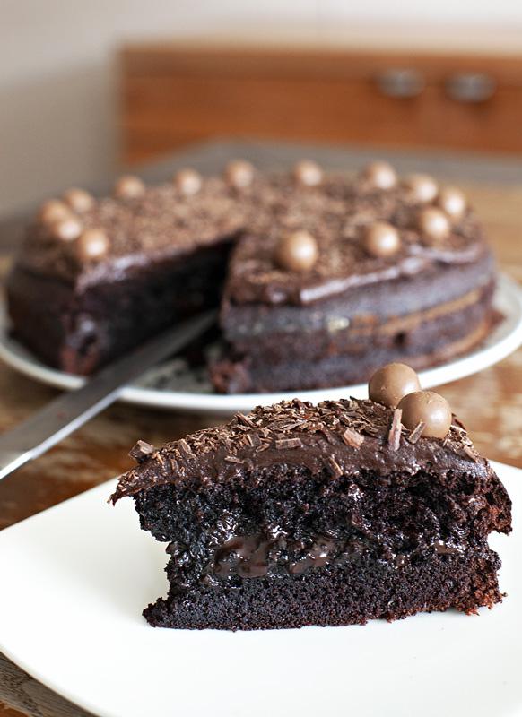 A slice of rich chocolate cake with chocolate frosting and chocolate balls on top sits on a white plate, embodying the irresistible charm of homemade desserts, with the rest of the cake in the background.