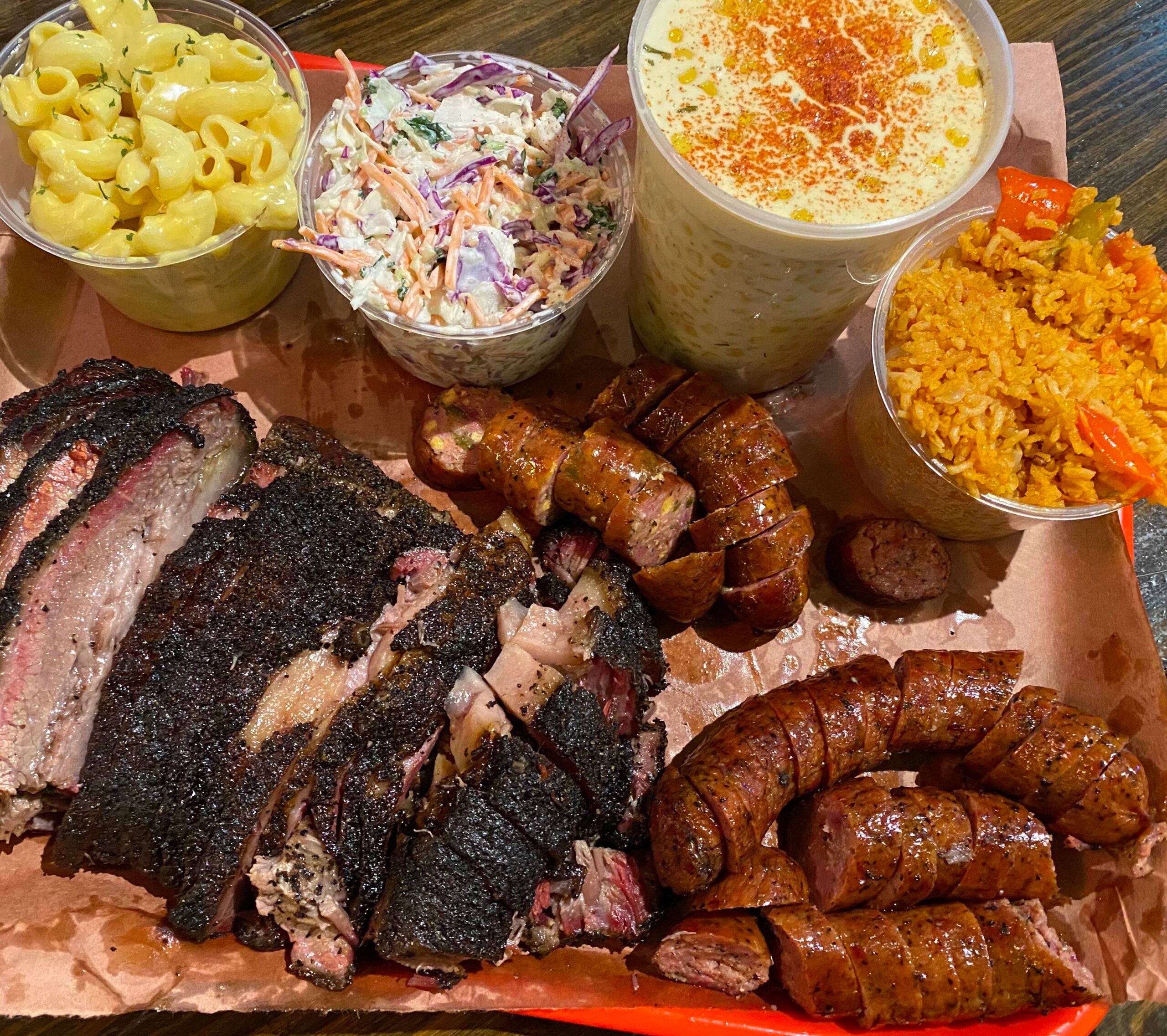 A tray with sliced brisket, BBQ sausage, macaroni and cheese, coleslaw, creamed corn, and seasoned rice with carrots in small cups, served on brown paper.