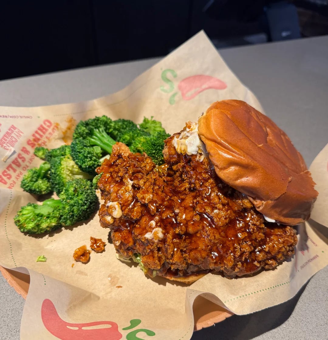 A fried chicken sandwich with a glazed, crispy patty sits open-faced on paper next to a side of steamed broccoli on a plate.