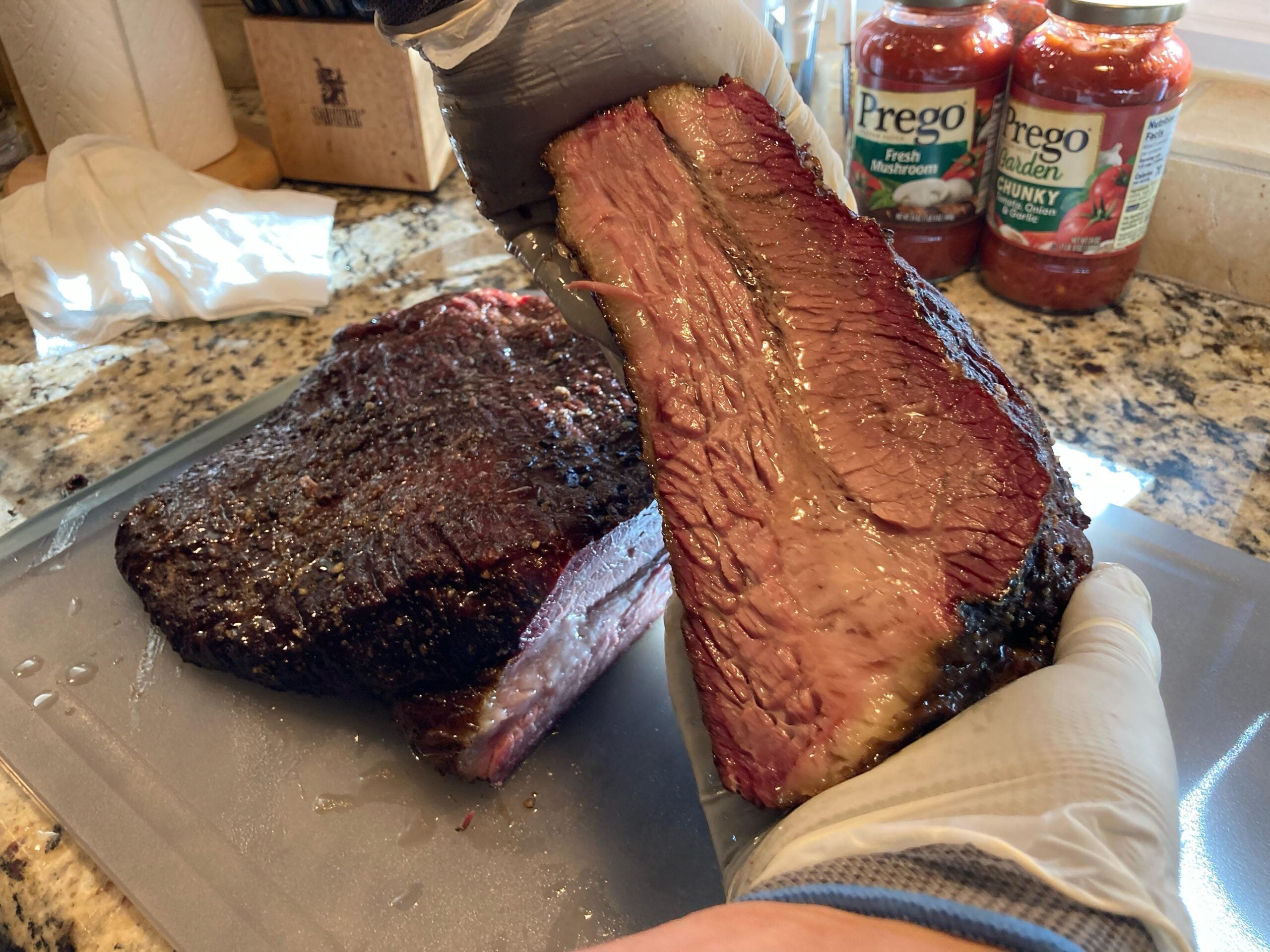 A gloved hand holds up a juicy, sliced smoked brisket on a cutting board, showing the pink smoke ring. Jars of Prego pasta sauce are visible in the background on a granite countertop.