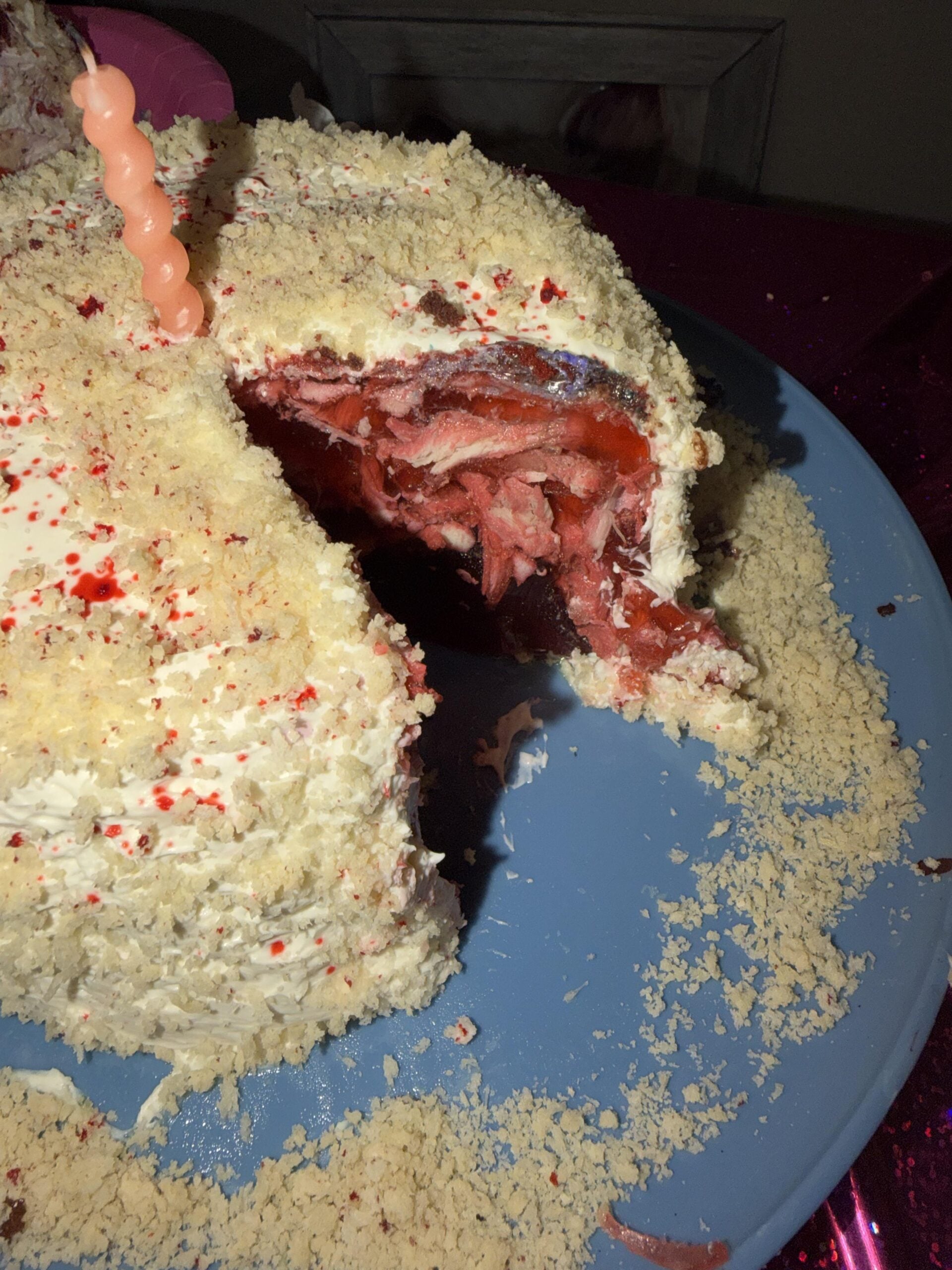 A cake with white frosting and crumbs sits on a blue plate. A large slice is cut out, revealing a red, jam-filled interior. There are crumbs scattered on the plate and a pink spiral candle on top.