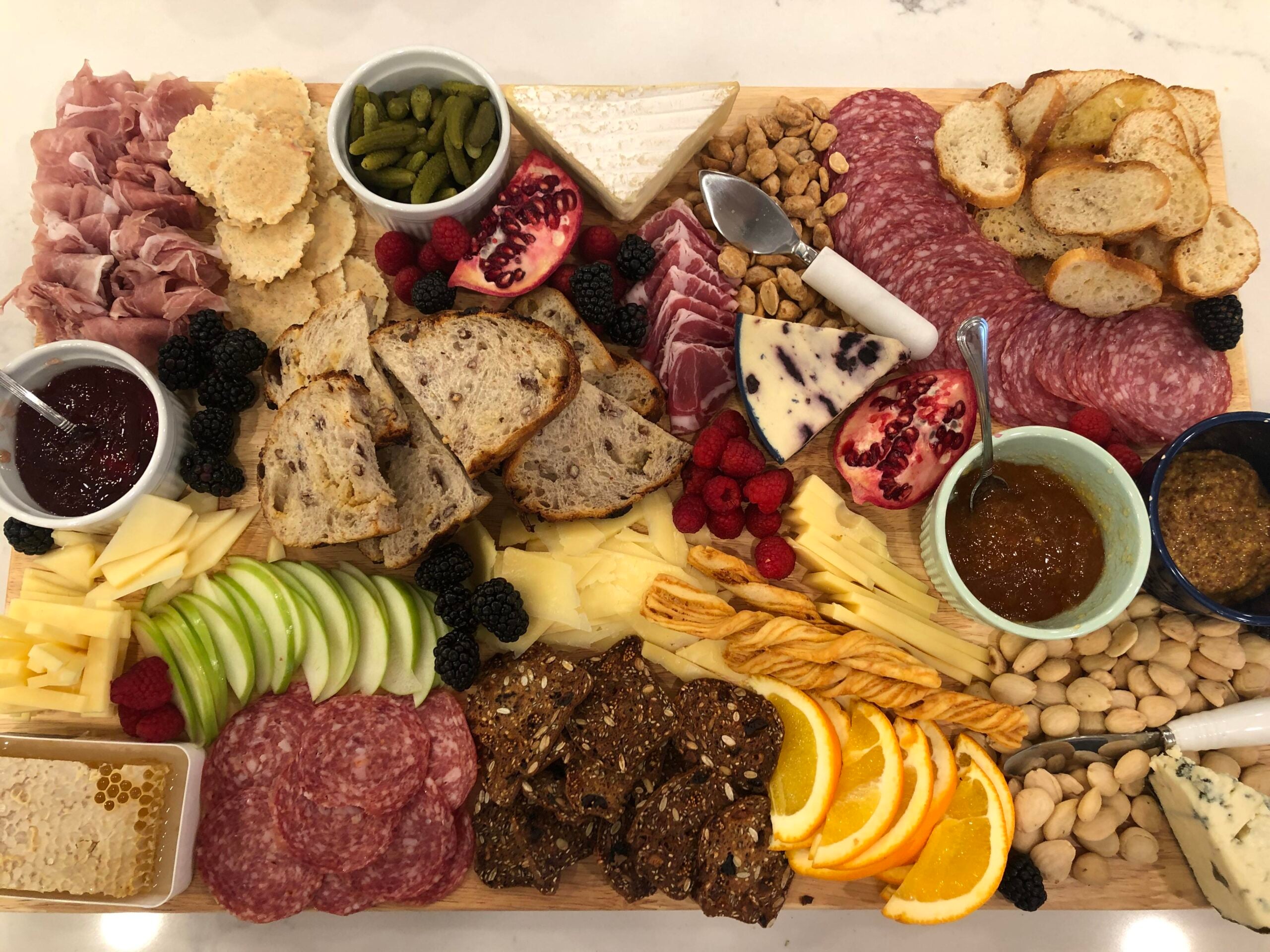 A charcuterie board with assorted cheeses, cured meats, bread slices, crackers, apple and orange slices, blackberries, pomegranate, jams, pickles, nuts, breadsticks, honeycomb, and spreads, arranged on a wooden board.