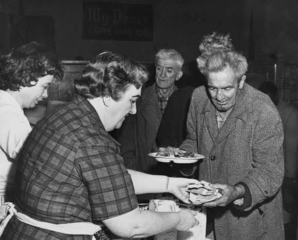 A woman serves food to an older man holding a plate in a soup kitchen, while other people wait in line. The scene, reminiscent of nostalgic dinners from the mid-20th century, captures a moment of warmth and community.