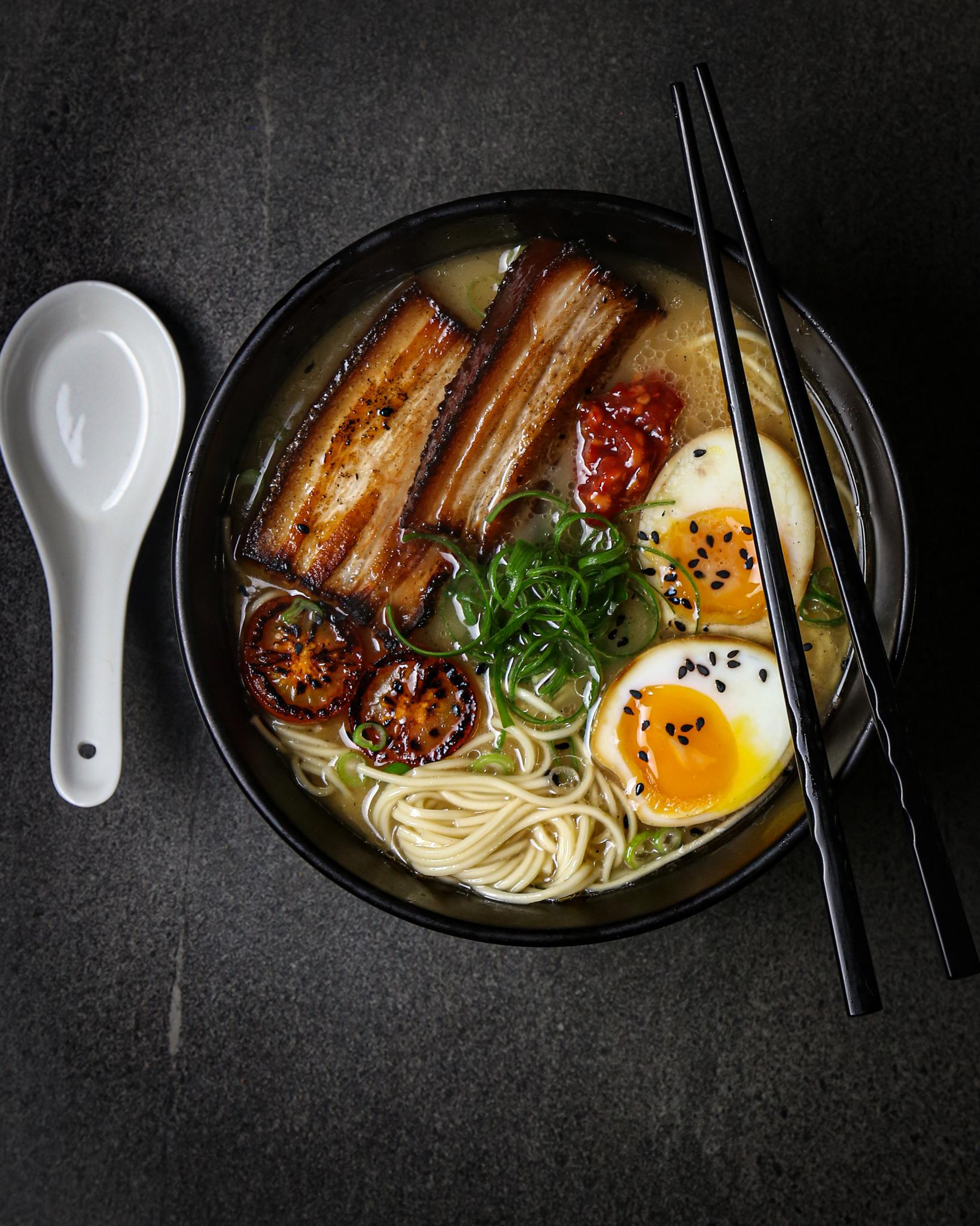 A bowl of ramen with noodles, sliced pork belly, soft-boiled eggs, grilled tomatoes, green onions, and chopsticks, set on a dark surface with a white soup spoon beside it.