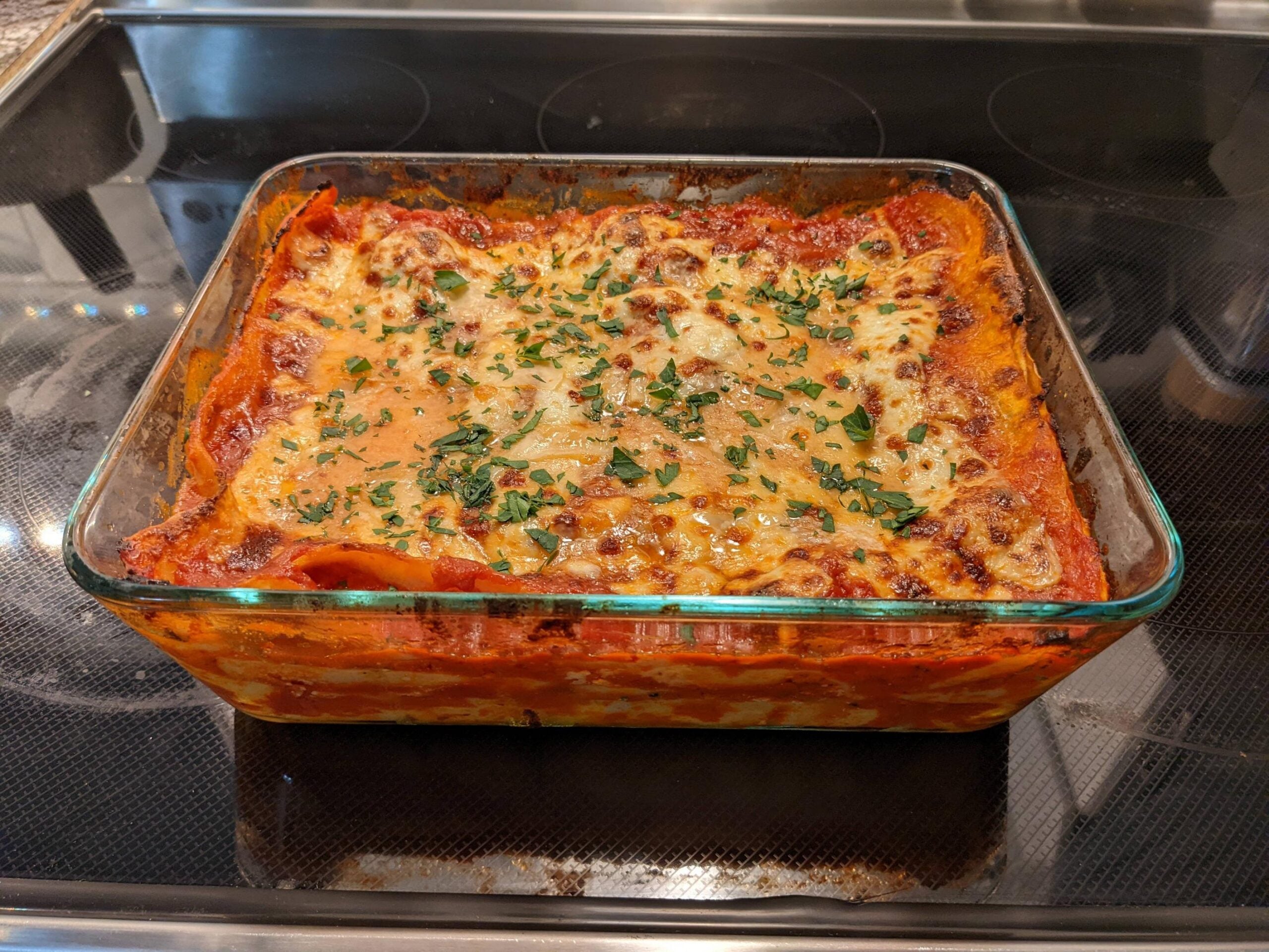 A glass baking dish filled with freshly baked lasagna, topped with melted cheese and chopped herbs, sits on a stove.