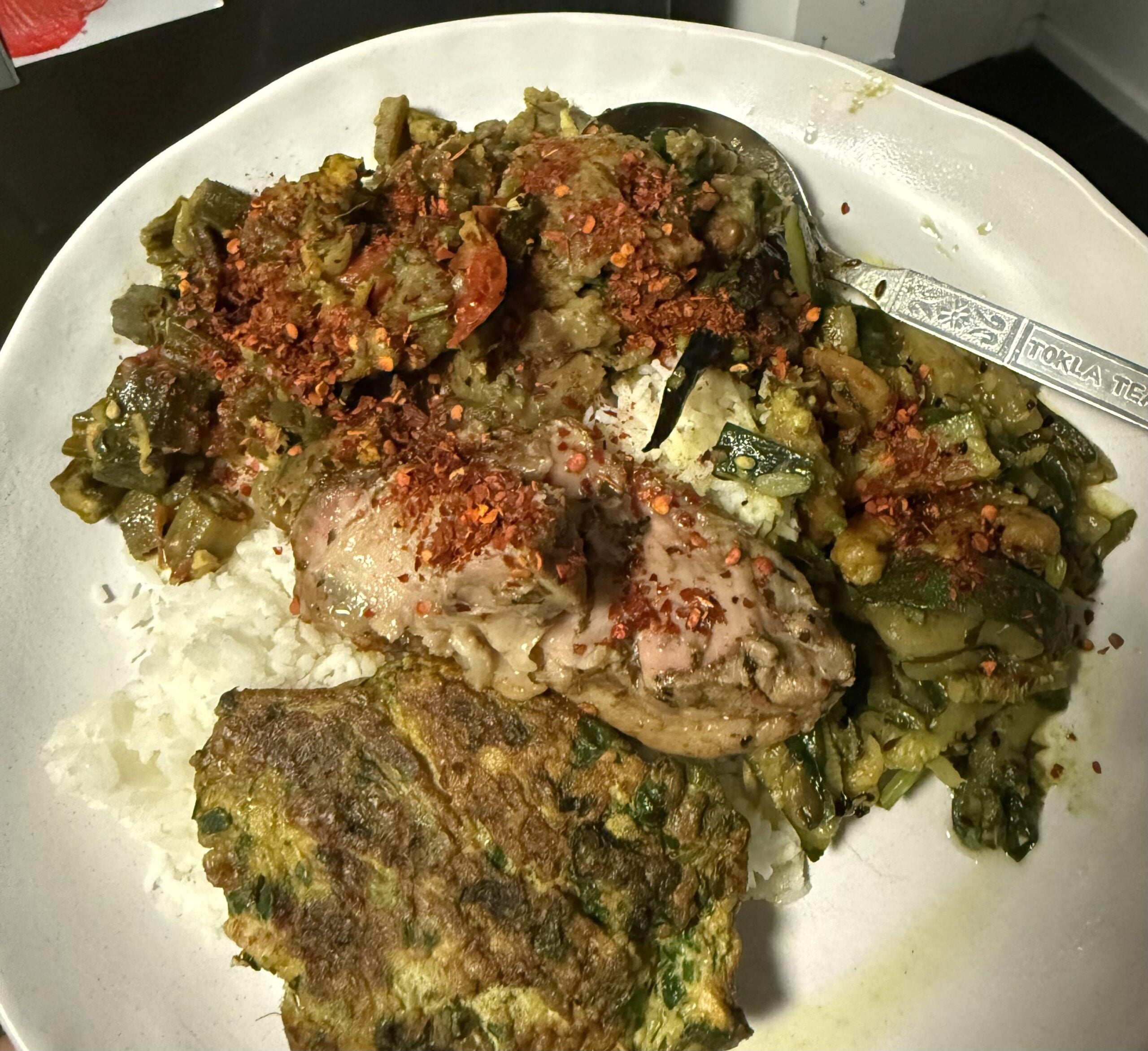 A plate of rice topped with cooked vegetables, a piece of seasoned chicken, sprinkled chili flakes, and a serving of herb omelette, with a spoon resting on the plate.