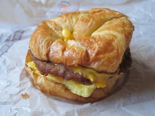 A breakfast sandwich with sausage, egg, and melted cheese inside a flaky croissant, placed on a crumpled wrapper.