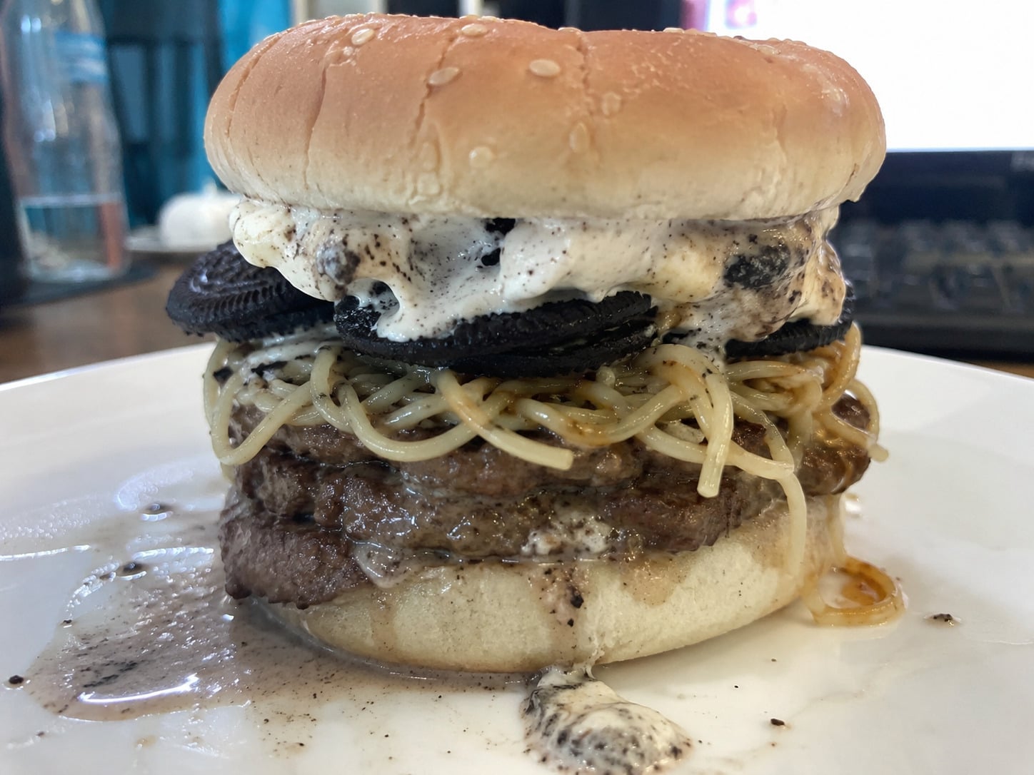 A burger with ground beef, spaghetti noodles, Oreos, and melted ice cream on a bun, sitting on a white plate.