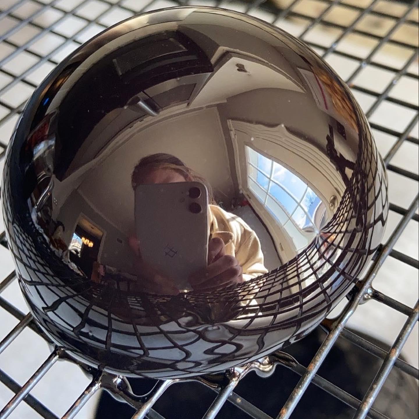 A shiny, dark, round object sits on a cooling rack, reflecting a person taking a photo with a smartphone and a window with multiple panes.