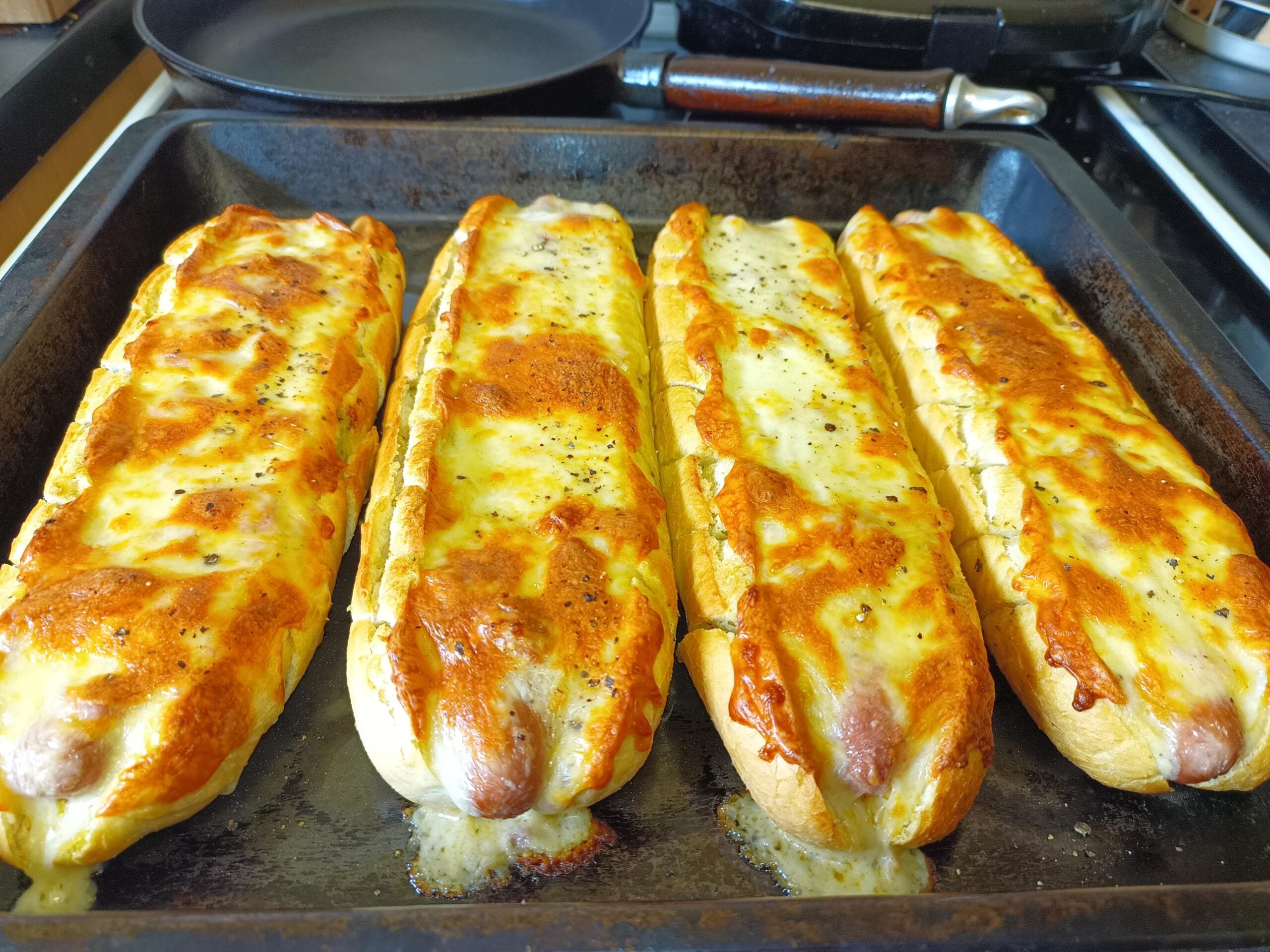 Four baked cheese-covered hot dogs in buns are lined up on a baking tray, golden and melted on top, with pepper sprinkled over the cheese. The tray is in a kitchen setting.