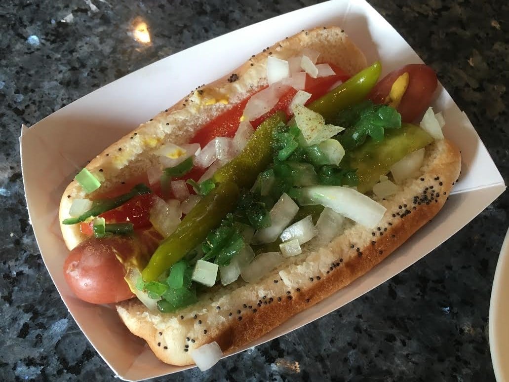 A Chicago-style hot dog in a poppy seed bun, topped with mustard, relish, onions, tomato, sport peppers, and a pickle spear, served in a paper tray.