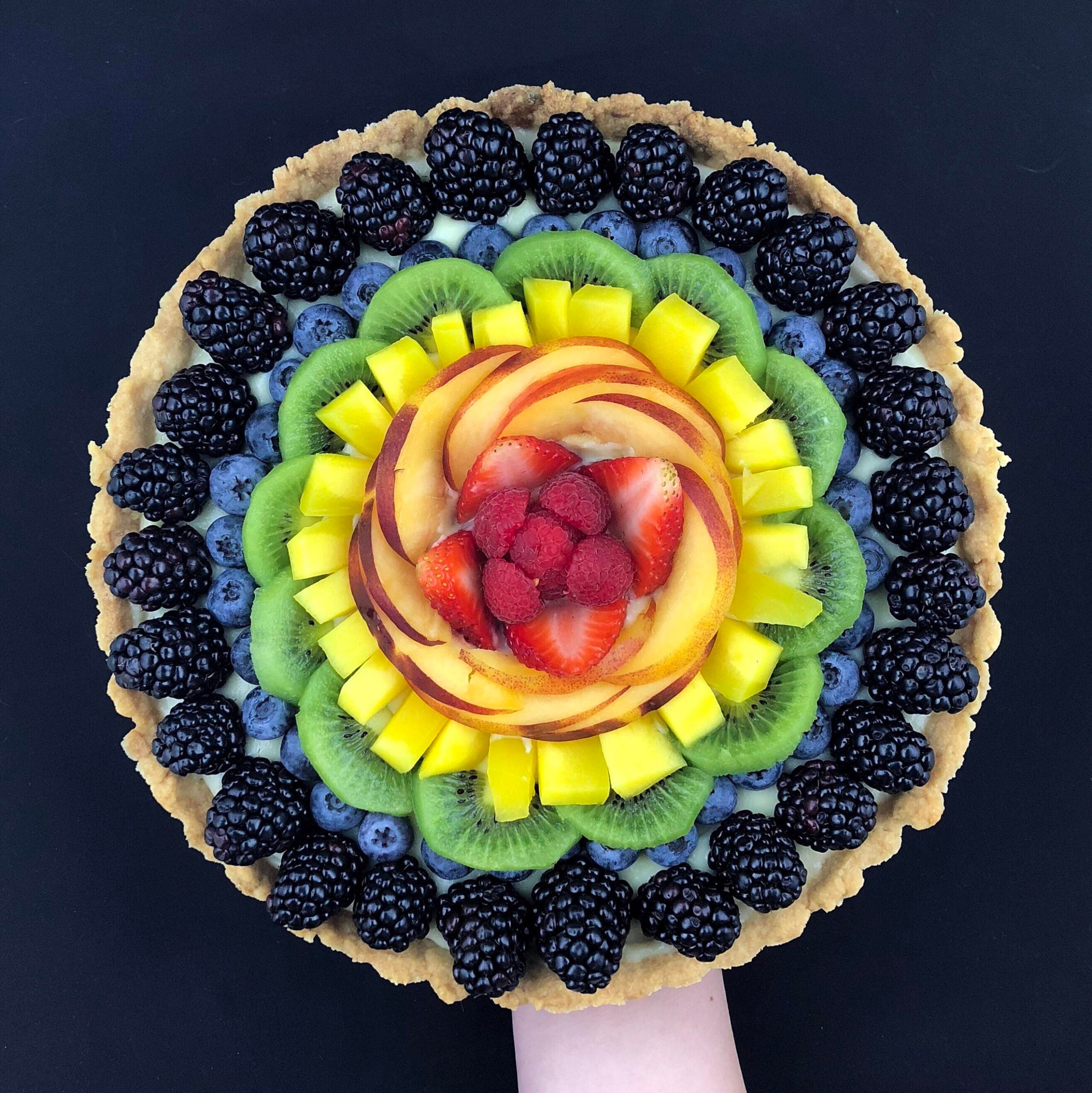 A colorful fruit tart topped with blackberries, blueberries, kiwi, mango, peach slices, strawberries, and raspberries, arranged in concentric circles on a black background.