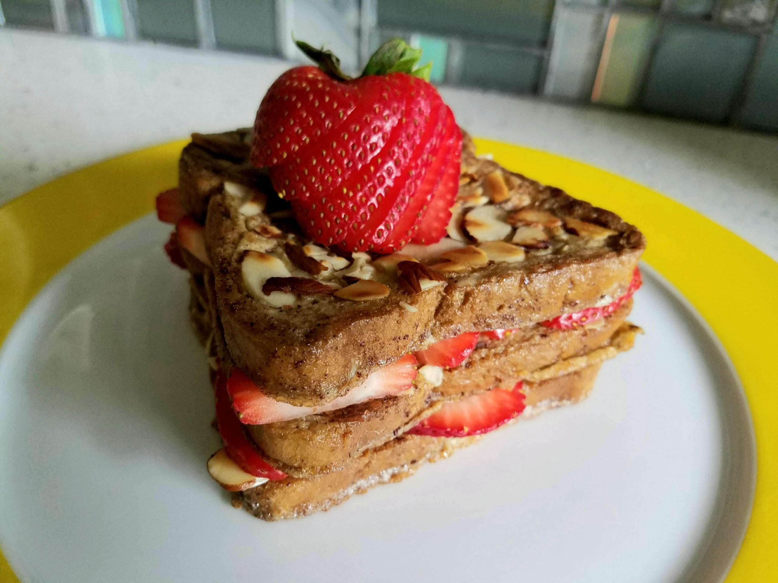 A stack of heart-shaped French toast with sliced strawberries and almond slivers, topped with a whole strawberry, on a white plate with a yellow rim.