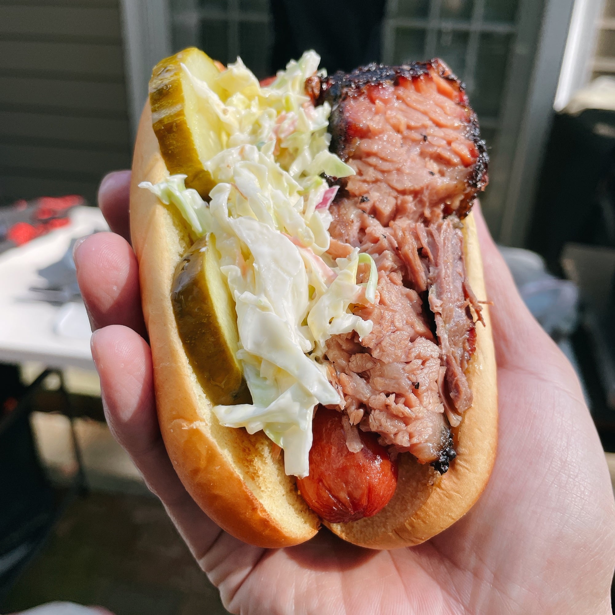 A hand holds a hot dog topped with shredded barbecue brisket, coleslaw, and pickle slices in a bun. Outdoor patio scene is blurred in the background.