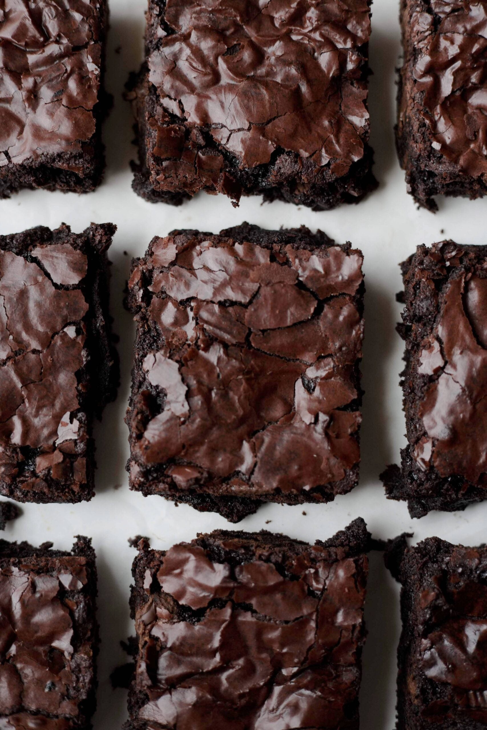 Nine chocolate brownies with shiny, cracked tops arranged in a grid pattern on a white surface.