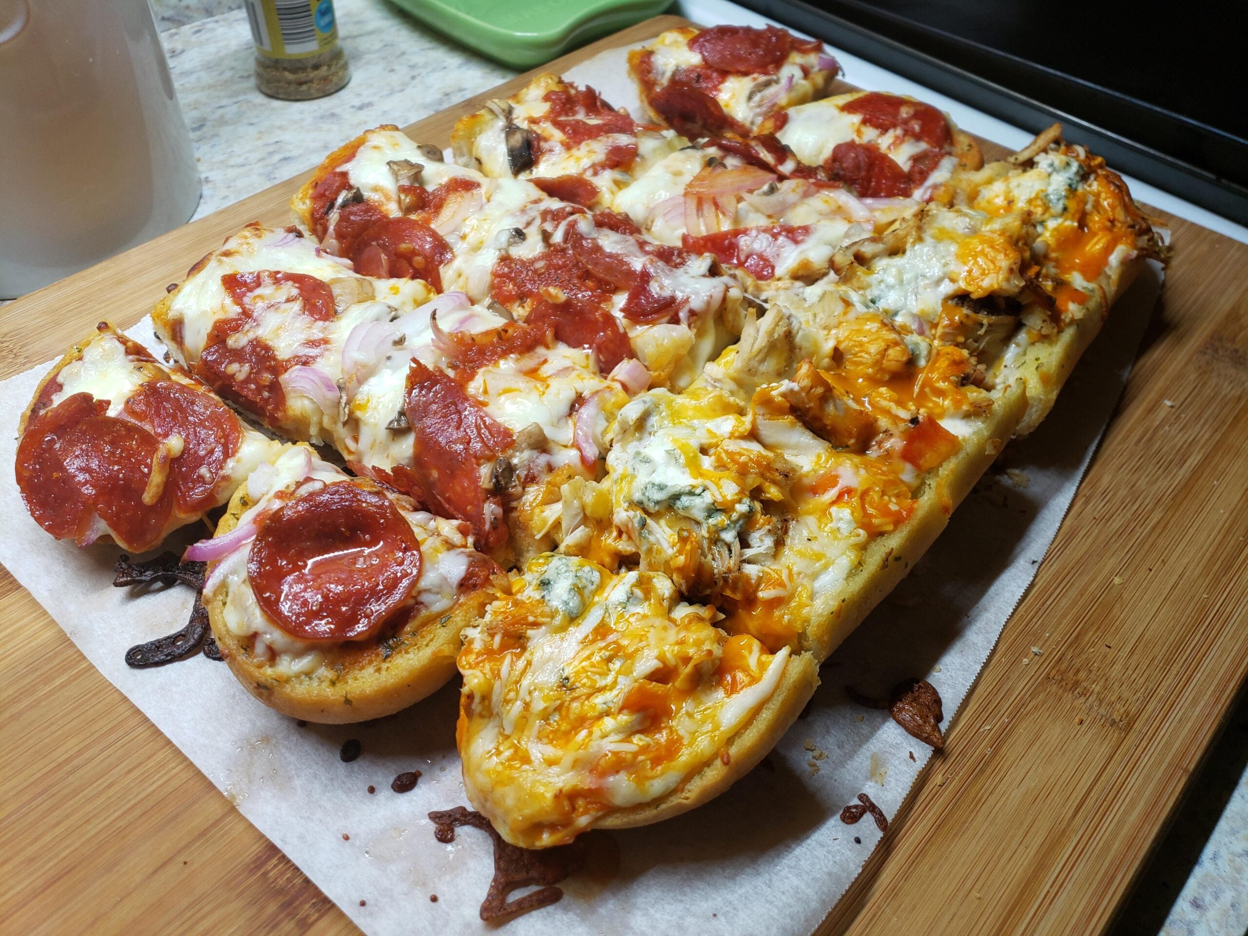 Rectangular homemade pizza on parchment paper, topped with pepperoni, onions, melted cheese, and herbs, cut into square slices and placed on a wooden cutting board.