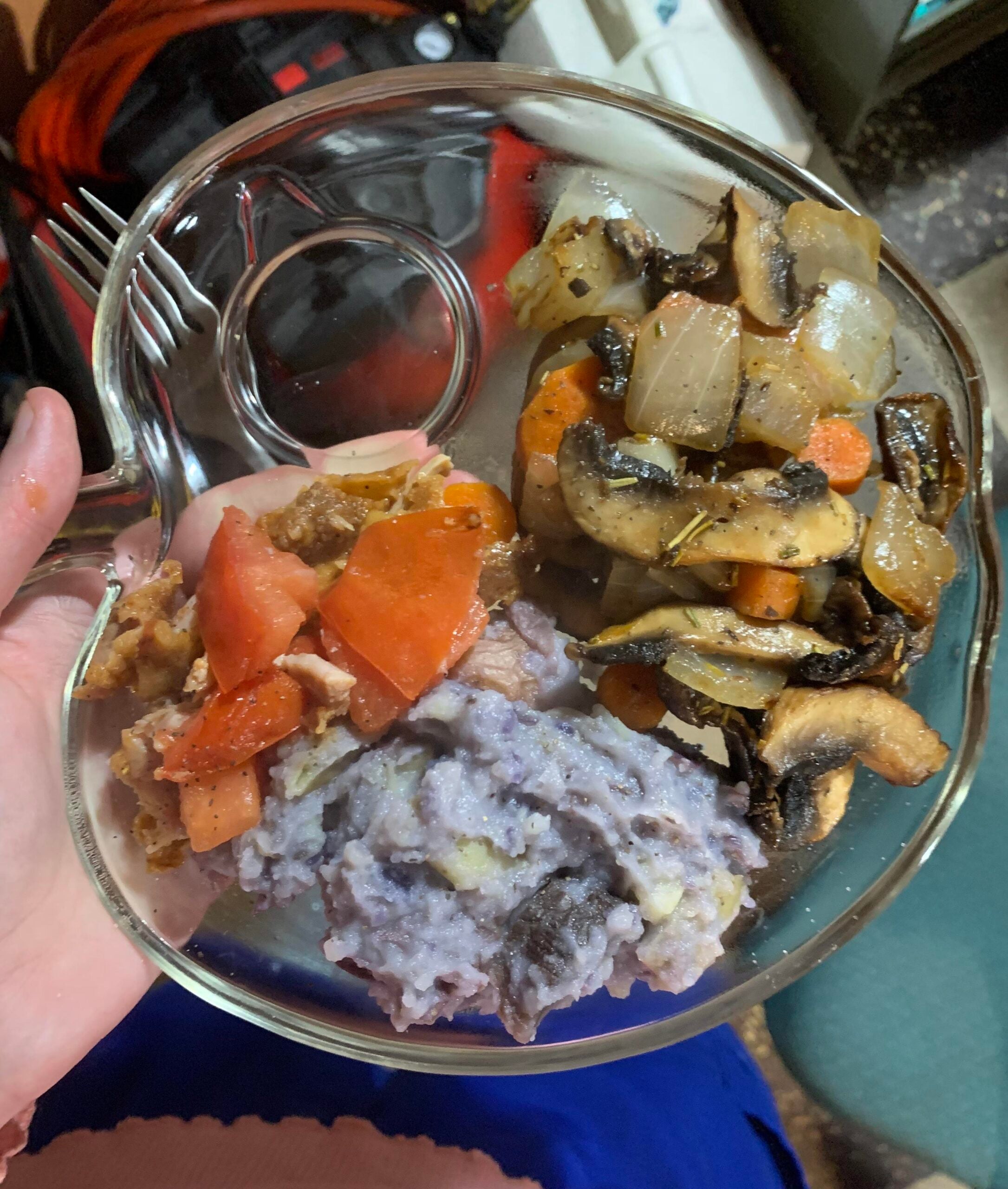 A clear plate holds purple mashed potatoes, roasted vegetables including mushrooms, onions, and carrots, and a tomato-based side. A fork rests on the plate.