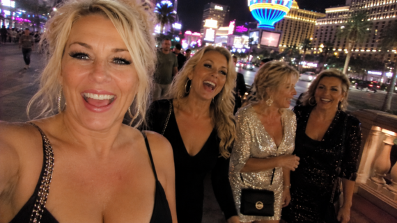 Four women dressed in glamorous evening wear are smiling and laughing while walking past neon lights and palm trees, with the dazzling backdrop of Las Vegas hotels illuminating the city street at night.