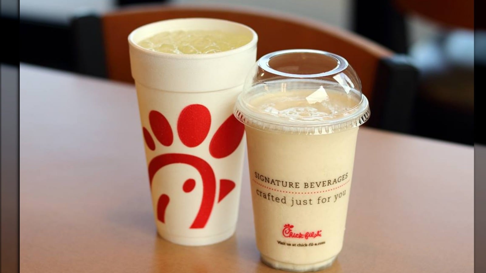 A large white cup with ice and a red logo sits next to a clear plastic cup of a creamy drink labeled “signature beverages” on a table. Both cups are from Chick-fil-A.
