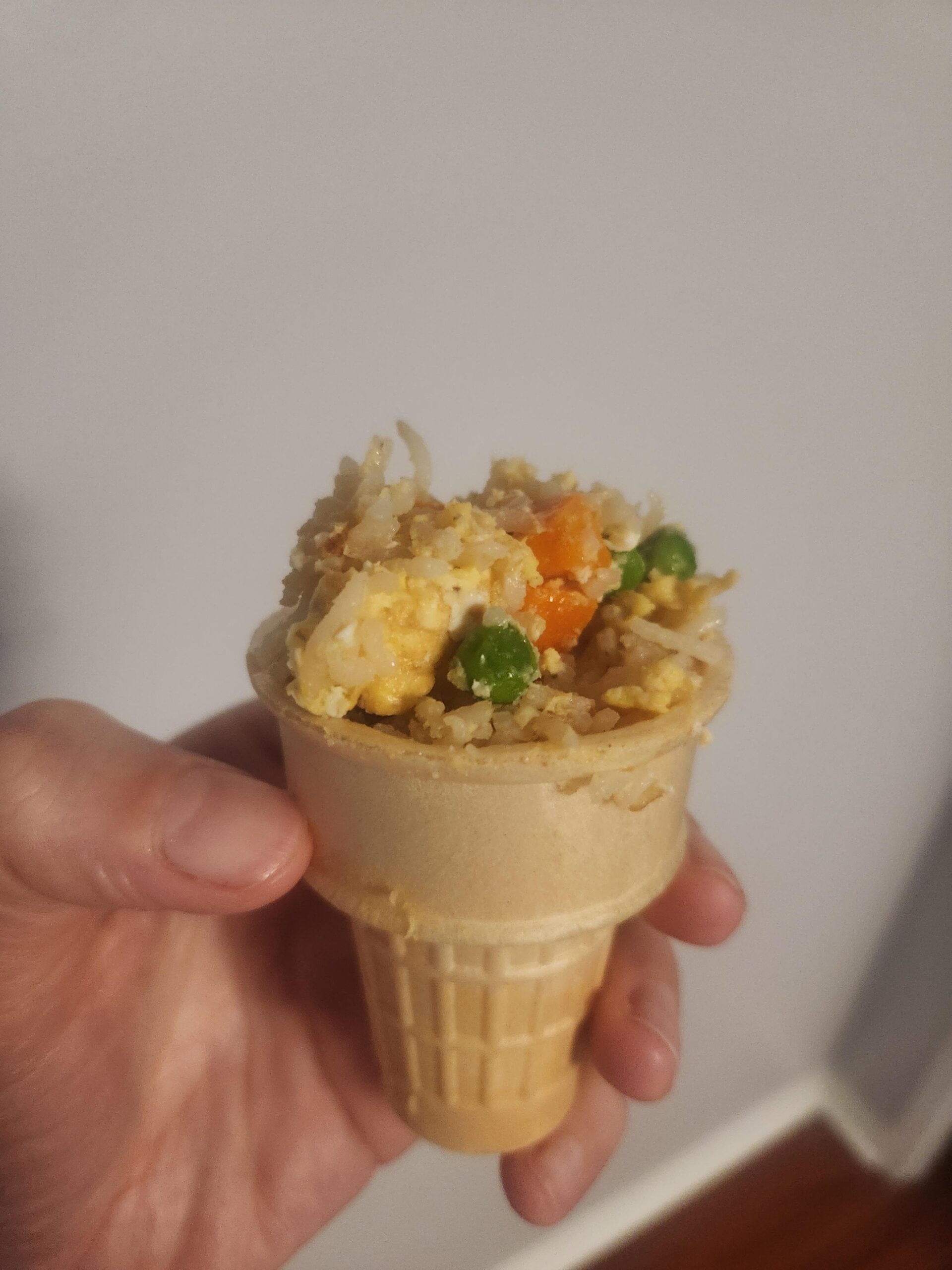A hand holding an ice cream cone filled with fried rice, peas, carrots, and scrambled eggs against a plain light-colored wall.