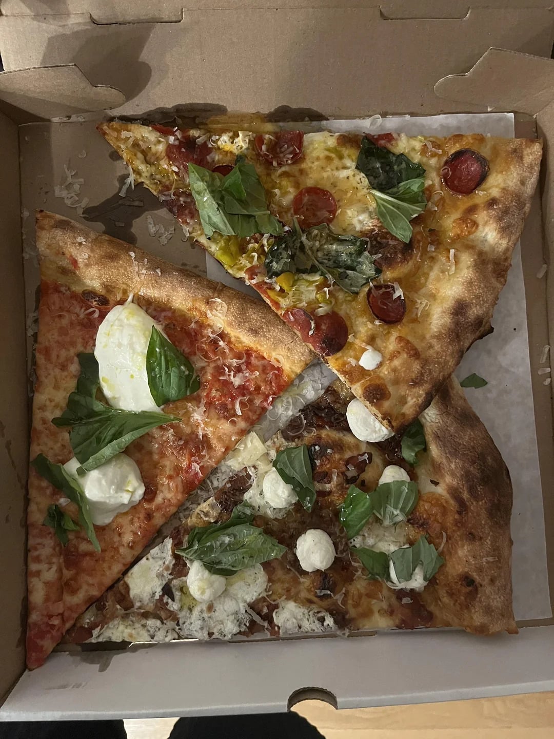 Three large pizza slices in a cardboard box, each with different toppings including basil leaves, pepperoni, cheese, and vegetables, sitting on parchment paper.