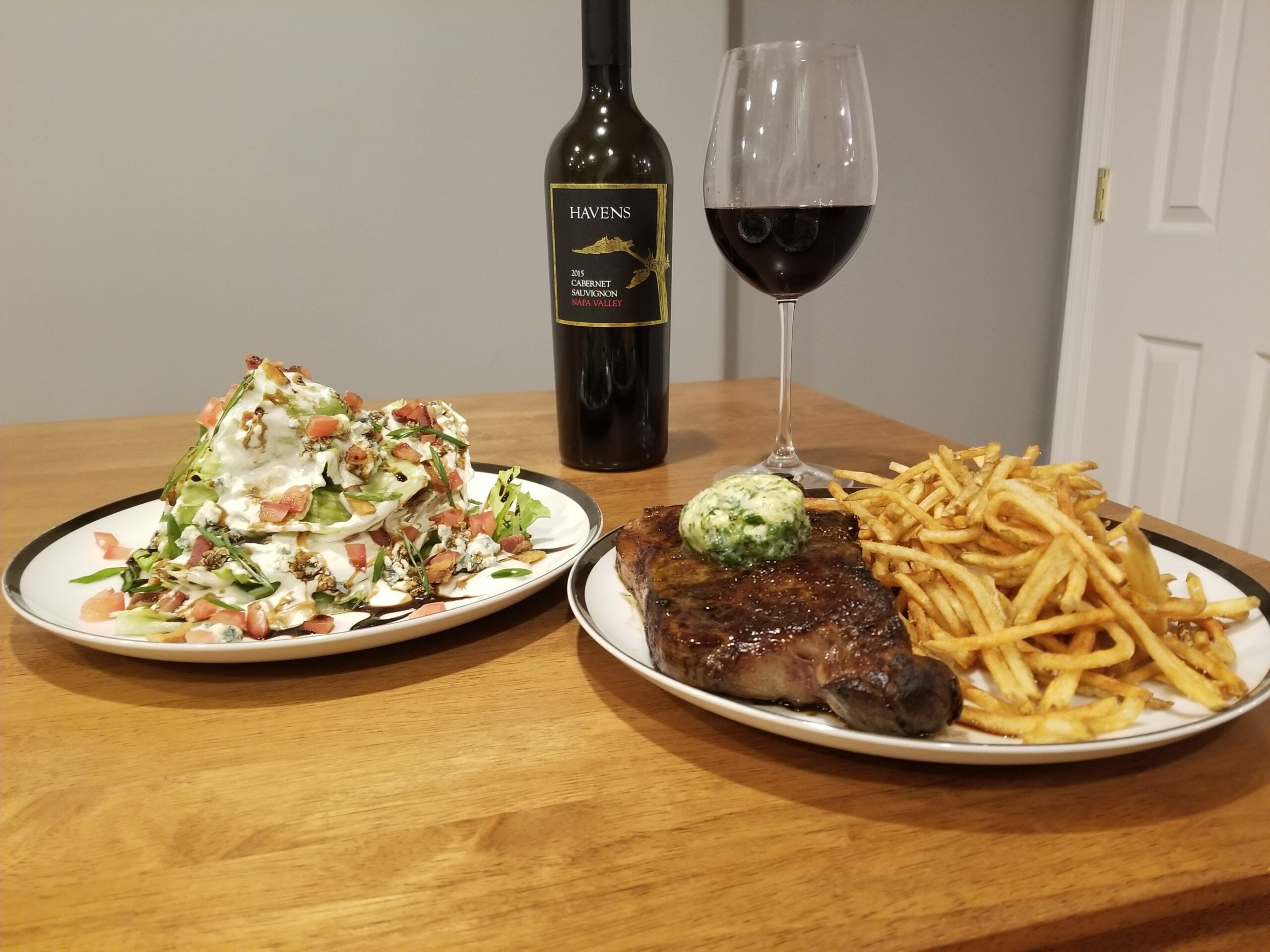A steak topped with herb butter, fries, and a wedge salad with tomatoes and dressing are on plates. A glass and bottle of red wine are on the wooden table.