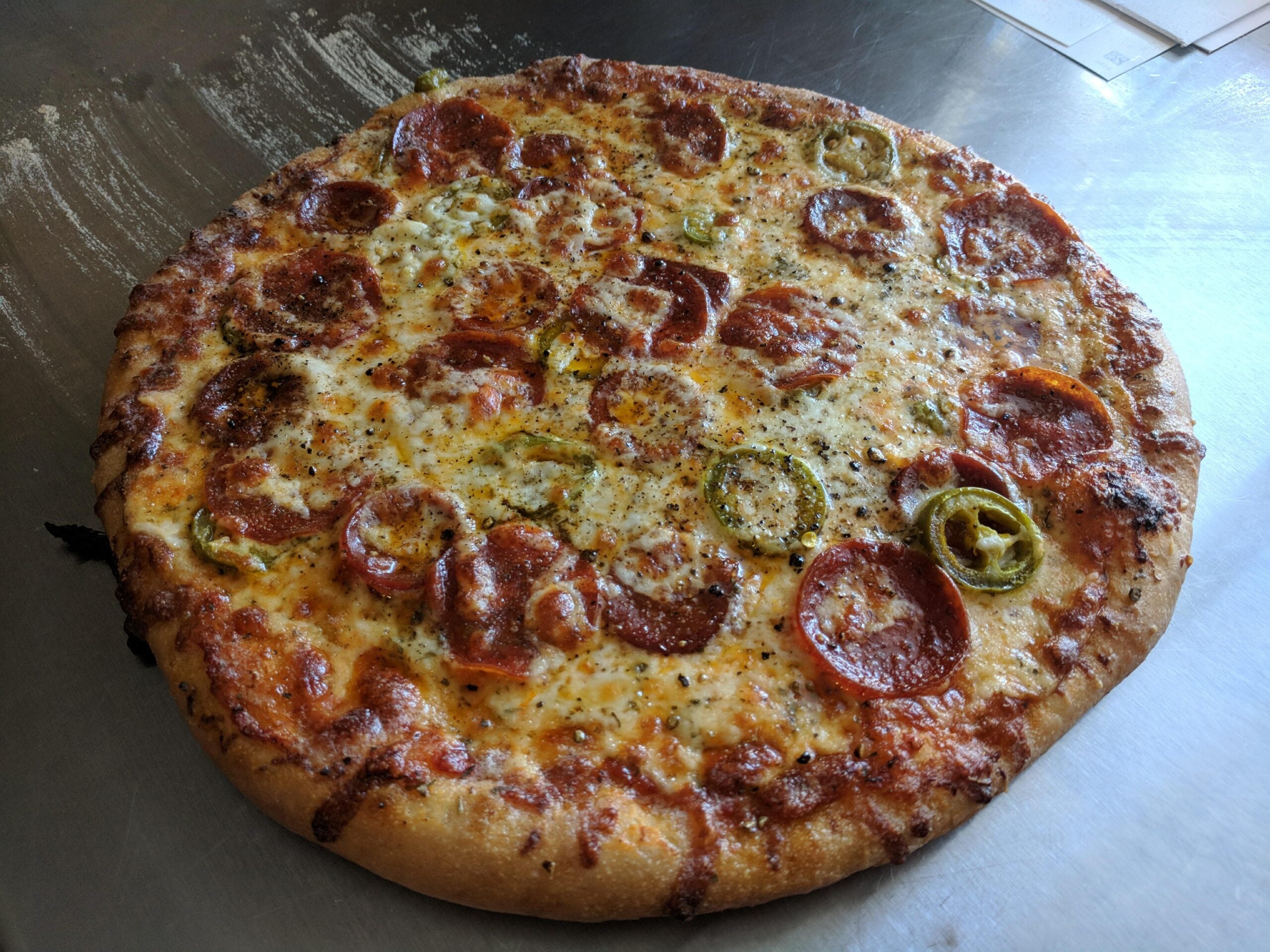A whole pepperoni pizza topped with melted cheese and slices of green and yellow peppers on a metal surface.