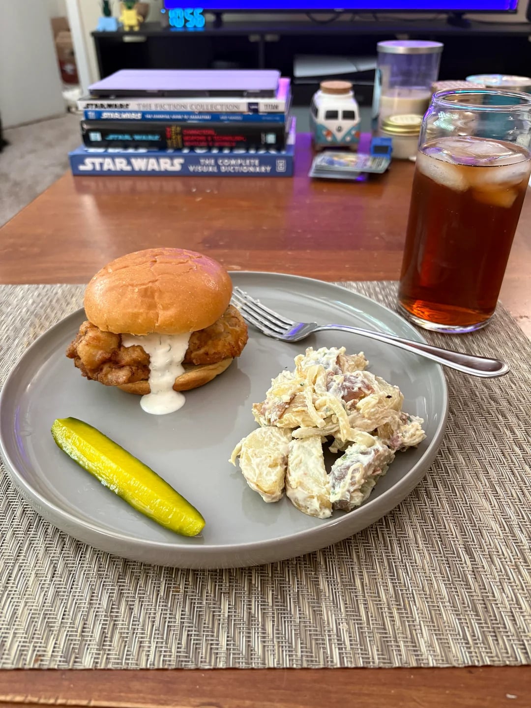 A plate with a chicken sandwich, potato salad, and a pickle spear sits on a placemat. A glass of iced tea, a fork, and books are on the table in the background.