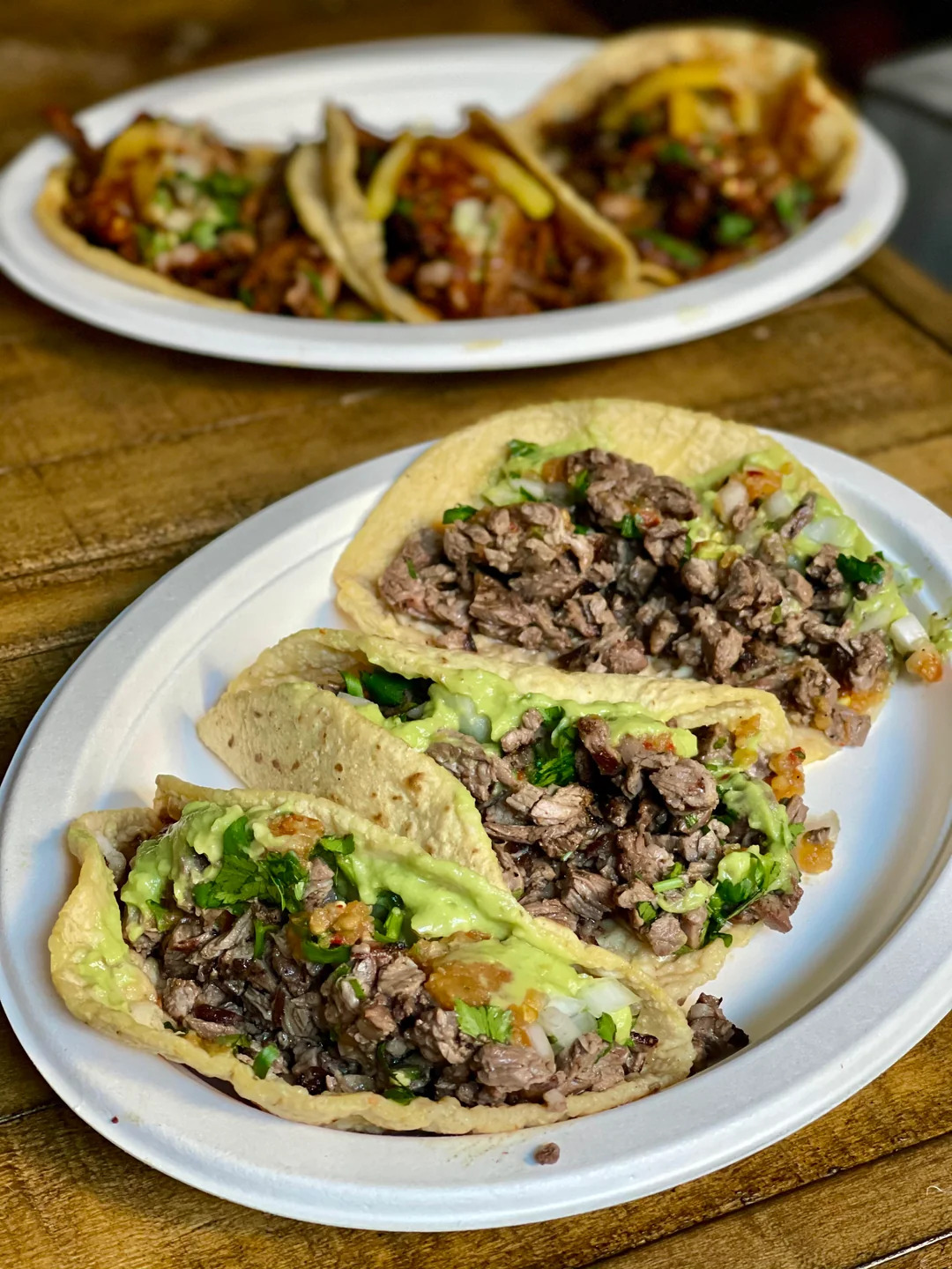 Three steak tacos topped with guacamole, onions, and cilantro are served on a white plate in the foreground. In the background, another plate with different tacos is slightly out of focus. Both are on a wooden surface.