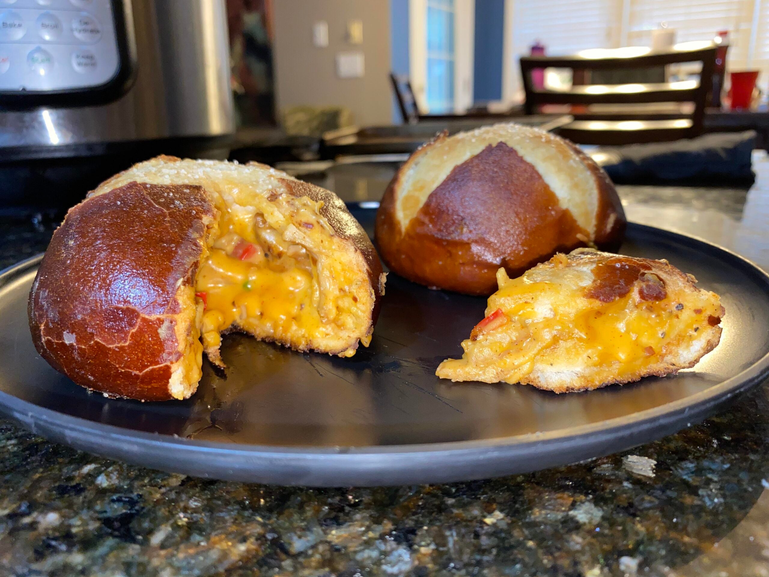 Three pretzel buns on a black plate, one sliced open to show a cheesy, orange filling with bits of red pepper. The plate sits on a dark countertop in a well-lit kitchen.