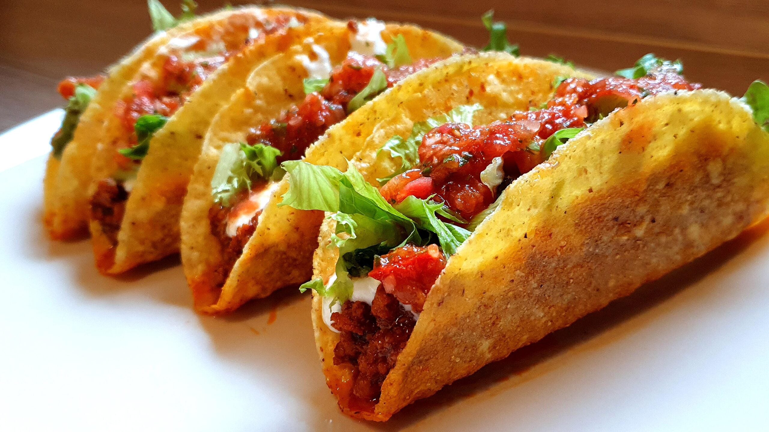 Four hard-shell tacos filled with ground beef, lettuce, tomato salsa, and cheese are arranged in a row on a white plate.