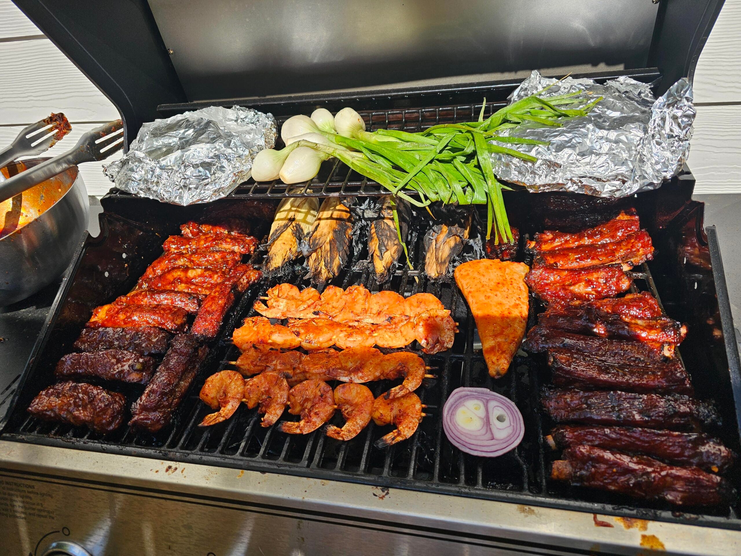 A grill loaded with skewered shrimp, ribs, corn, grilled vegetables, green onions, onions, foil-wrapped food, and round white vegetables, all cooking over open flames.