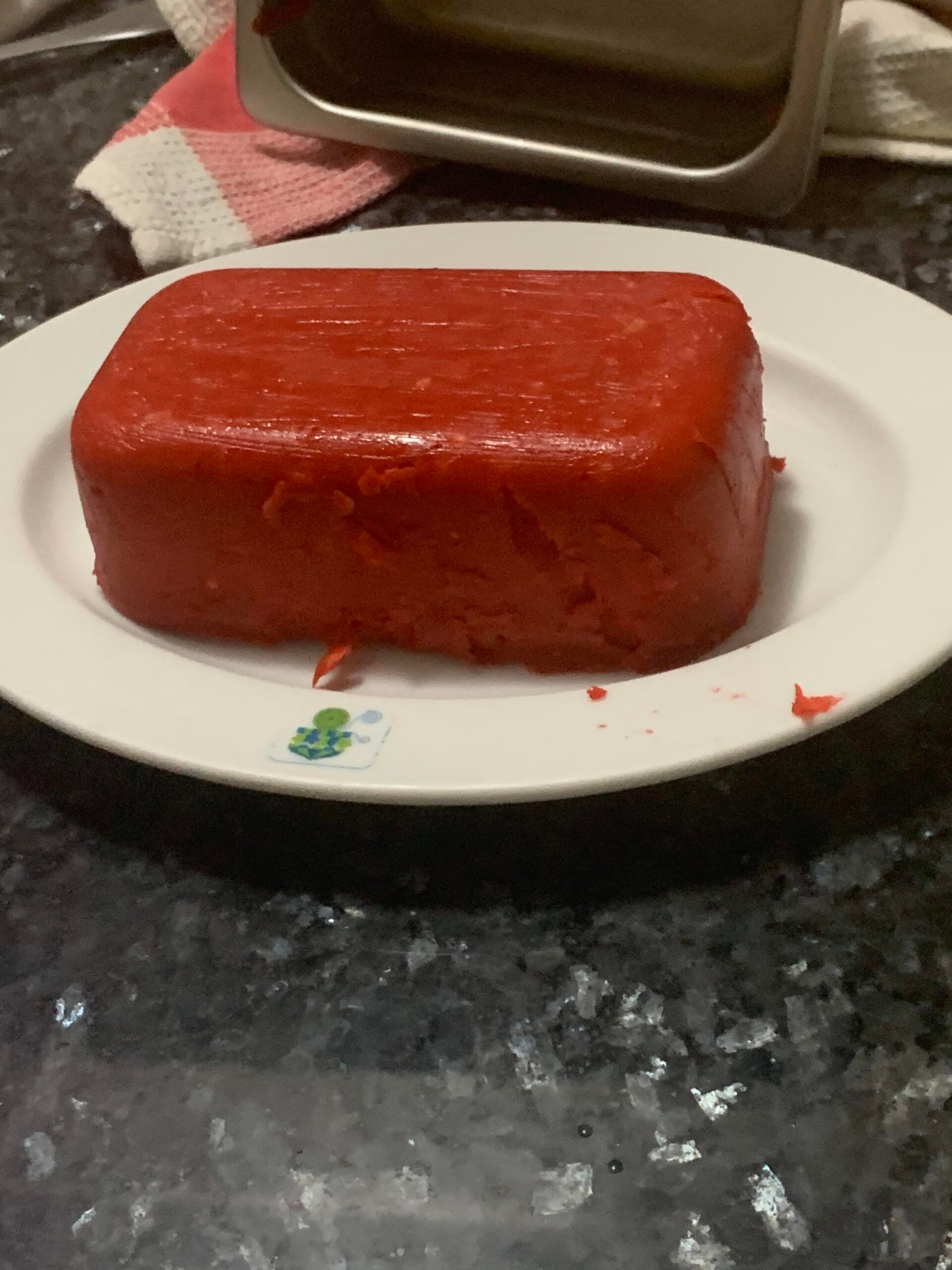 A rectangular red gelatin block sits on a white plate, placed on a dark countertop. Part of the gelatin appears to be scooped or cut out. A towel and metal container are visible in the background.