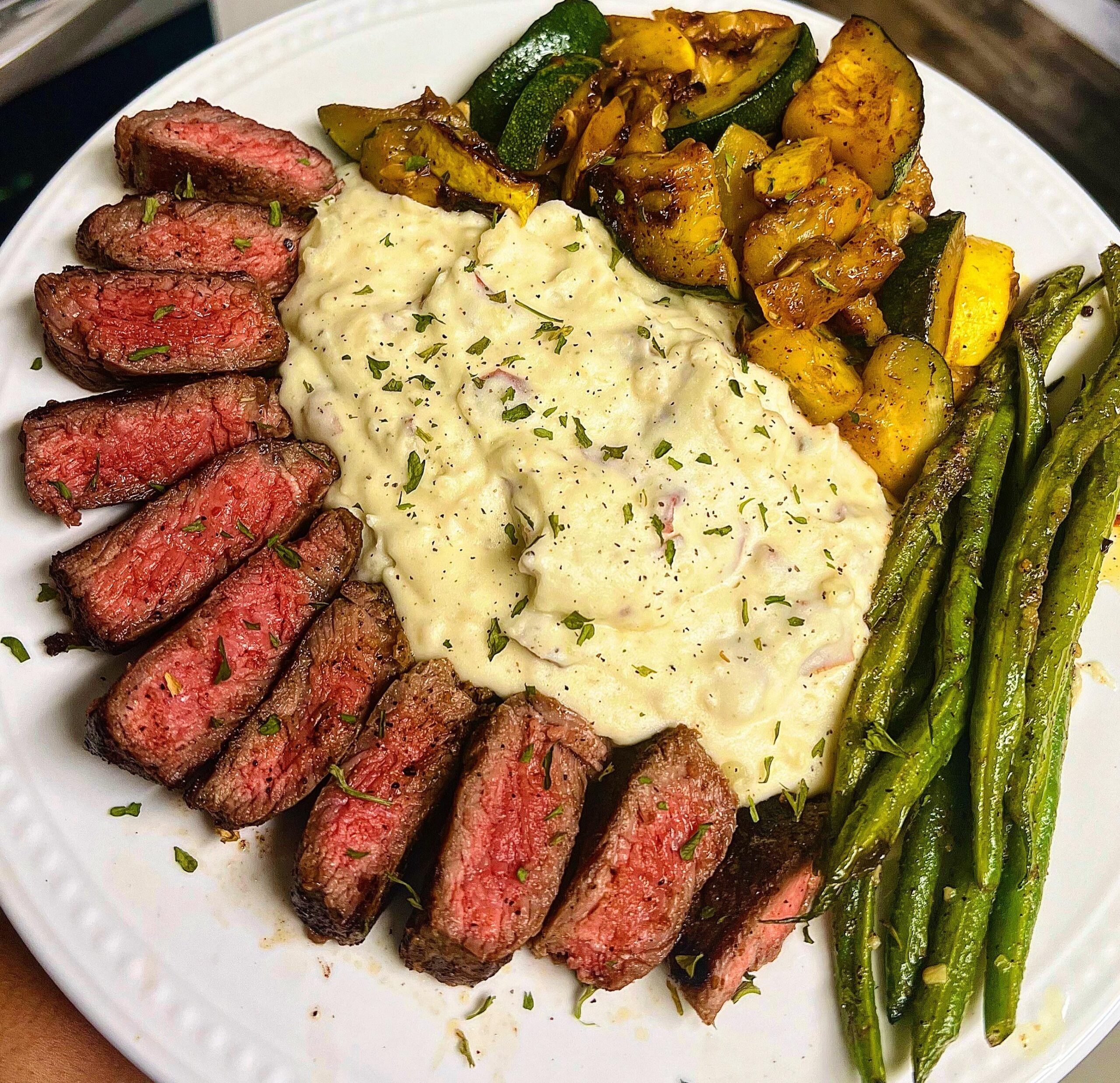 Sliced medium-rare steak, creamy mashed potatoes, sautéed zucchini and squash, and roasted green beans arranged on a white plate.