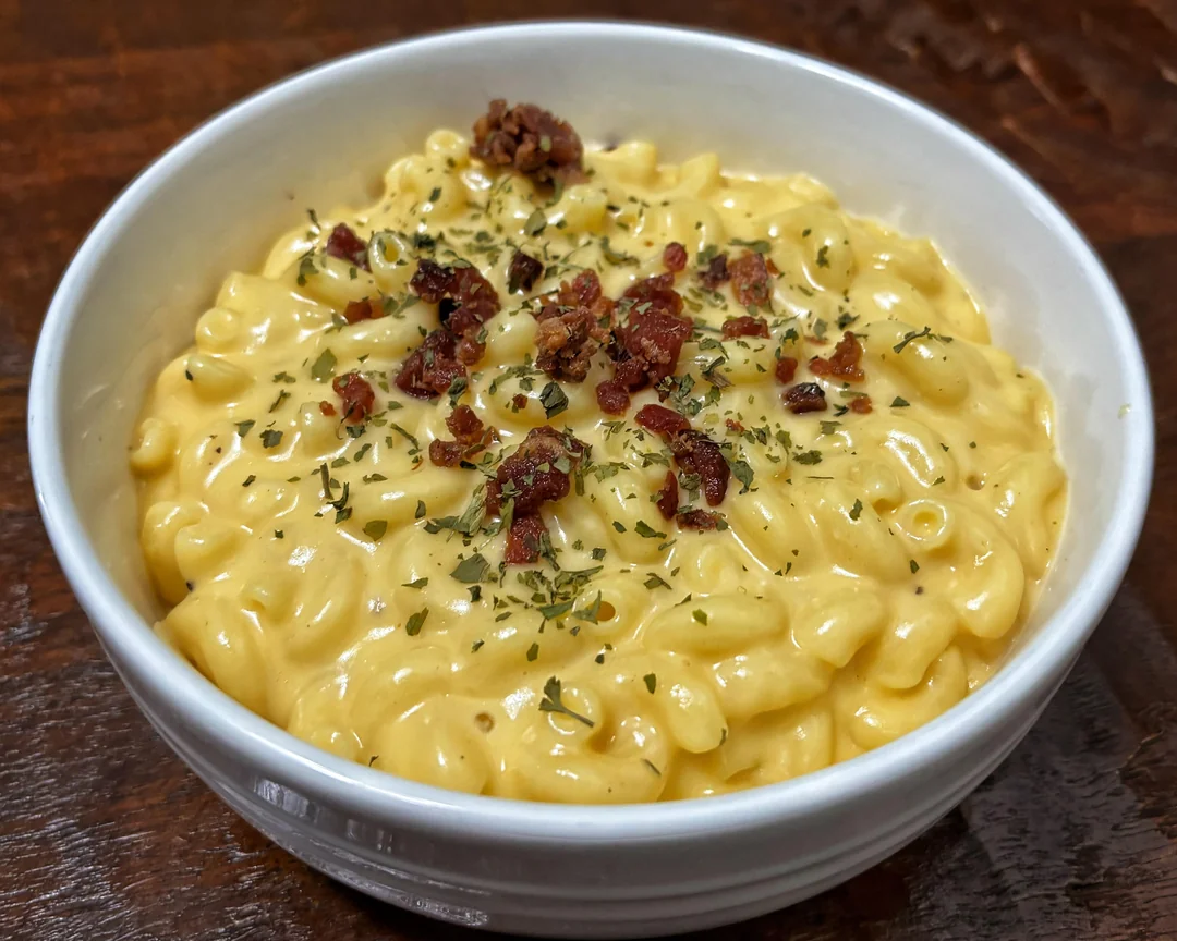 A white bowl filled with creamy macaroni and cheese, topped with crumbled bacon and sprinkled herbs, sits on a wooden surface—an inviting example of comforting homemade meals.