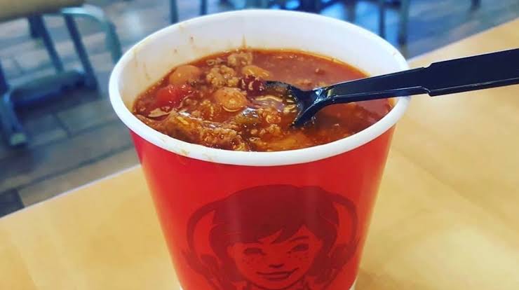 A red cup featuring the Wendy’s logo is filled with hearty chili containing ground beef and beans, with a black plastic spoon inside, placed on a light-colored table.