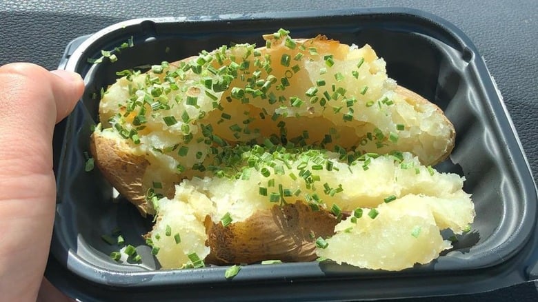 A hand holds a black plastic container with a baked potato split open and topped with chopped chives. The potato is fluffy inside and the skin is slightly crispy.