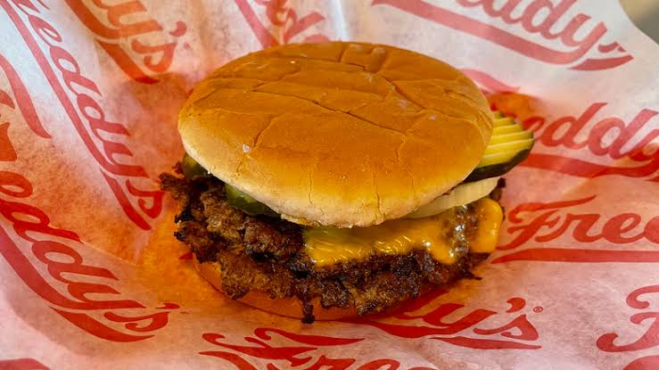 A cheeseburger with pickles on a branded Freddy's wrapper. The burger has a toasted bun, melted cheese, and a crispy beef patty.