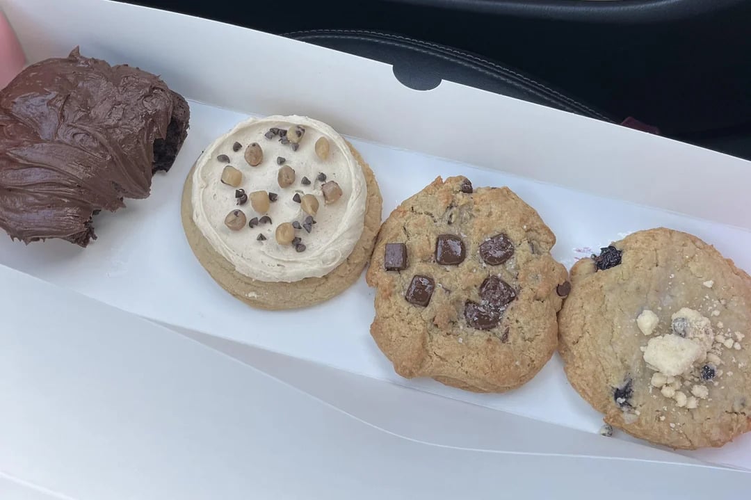 Four large cookies in a white box: one with thick chocolate frosting, one with white icing and chocolate chips, one with chocolate chunks, and one with blueberries and crumble topping—just like the irresistible treats you’d find among fast-food grocery store products.