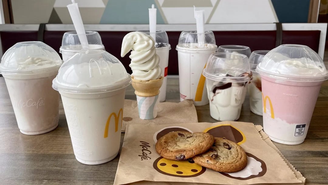 A variety of McDonald's desserts on a table, including milkshakes, an ice cream cone, sundaes, and two chocolate chip cookies on a McCafe napkin. The background shows a diner booth and patterned wall.