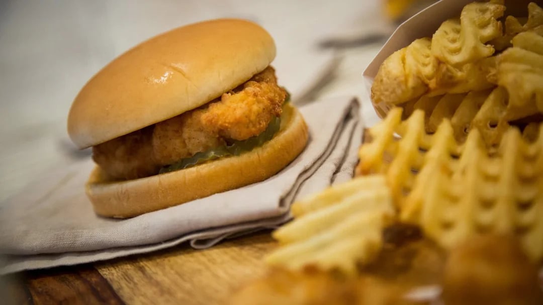 A chicken sandwich with pickles on a bun sits on a napkin next to a serving of waffle fries on a wooden surface.